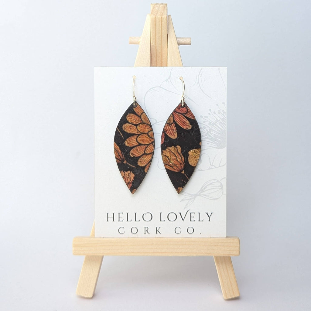 A pair of floral patterned earrings in autumn tones on a black background, displayed on a small wooden easel with the text 'Hello Lovely Cork Co.'.