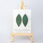 A pair of grass green cork earrings in a leaf shape, displayed on a small wooden easel with the brand name 'Hello Lovely Cork Co.'.