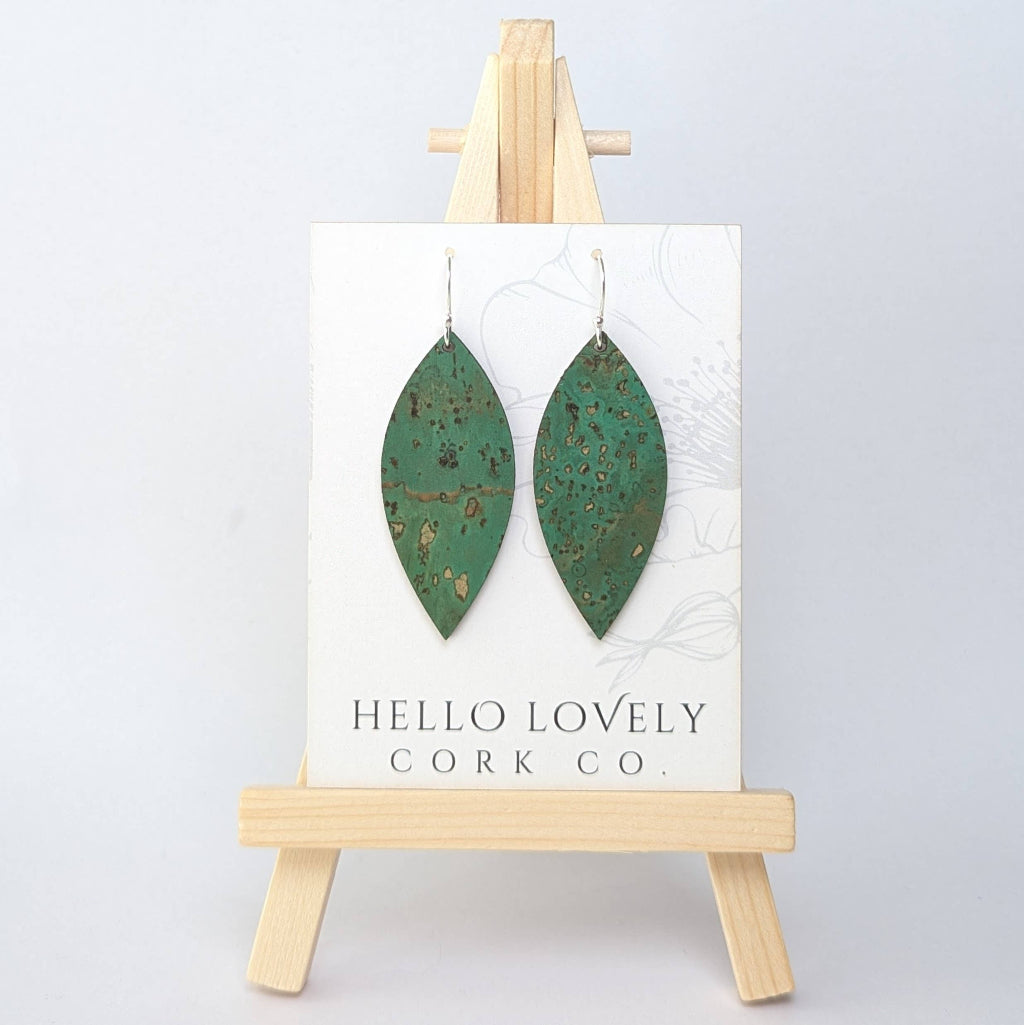 A pair of grass green cork earrings in a leaf shape, displayed on a small wooden easel with the brand name 'Hello Lovely Cork Co.'.
